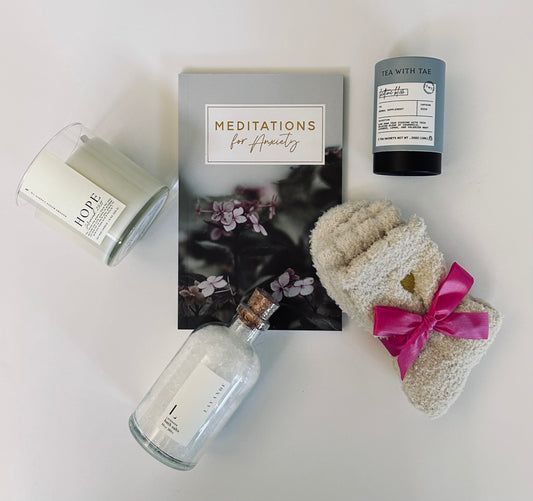 Anxiety and Peace Box contents laid out showing meditation for anxiety booklet, sleep tea tube, lavender bath salts jar, Hope candle with Hibiscus & Agave scent, and cozy socks for relaxation and self-care