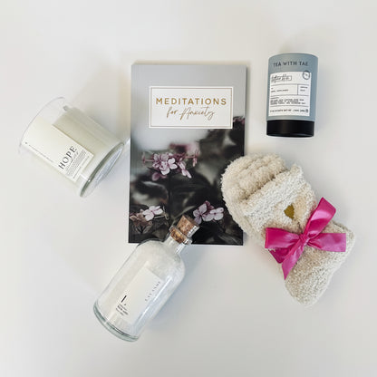 Anxiety and Peace Box contents laid out showing meditation for anxiety booklet, sleep tea tube, lavender bath salts jar, Hope candle with Hibiscus & Agave scent, and cozy socks for relaxation and self-care